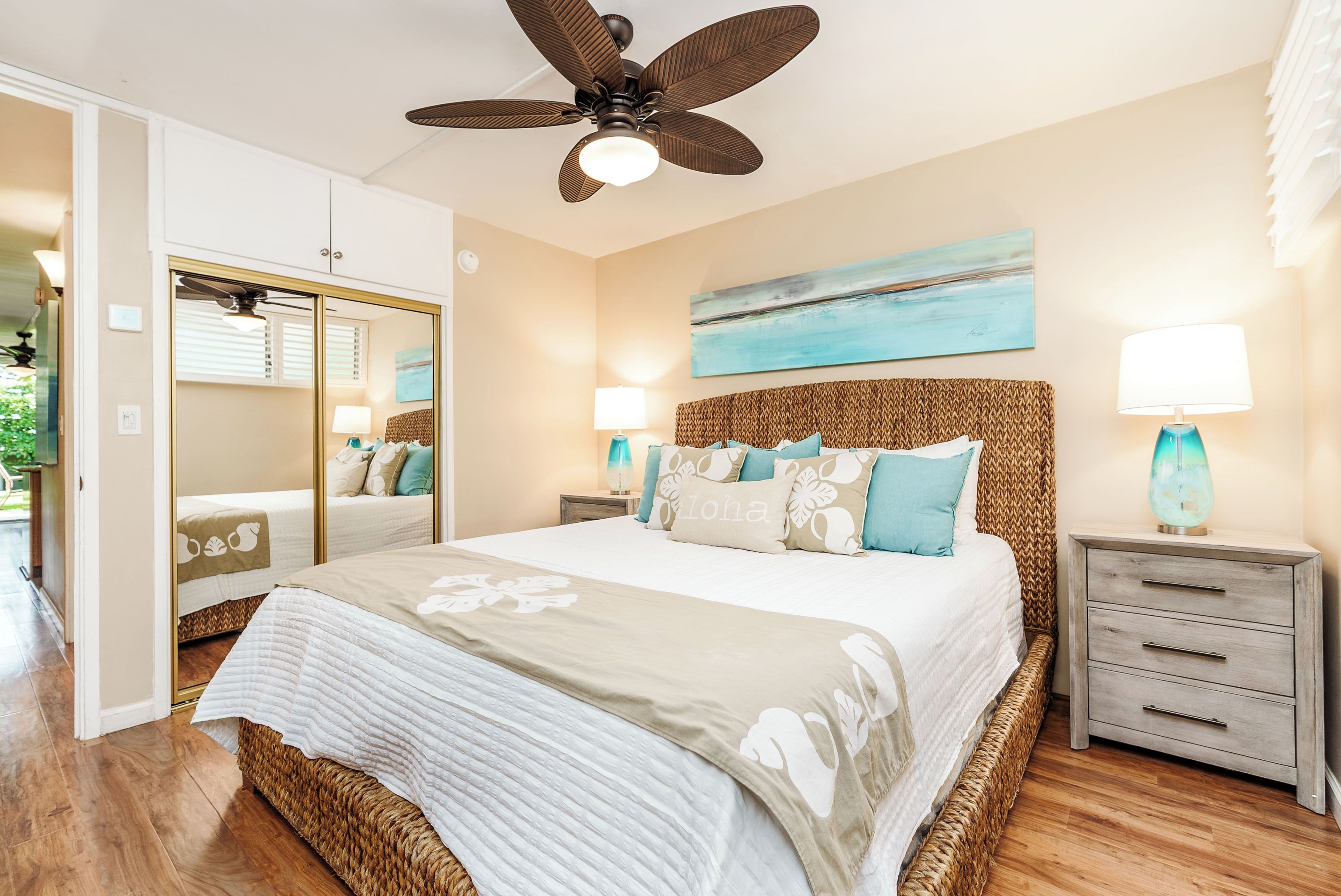 A bedroom with a large bed, a ceiling fan, a nightstand with a lamp, a wall-mounted painting, and a wardrobe with a mirror.