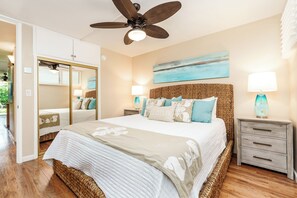 Une chambre avec un grand lit, un ventilateur de plafond, une table de chevet avec une lampe, un tableau accroché au mur et une armoire avec un miroir.