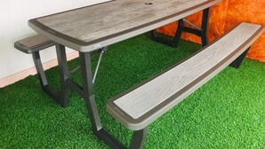 Una mesa de picnic con banco, sobre césped artificial, frente a una pared blanca con un panel naranja texturizado.