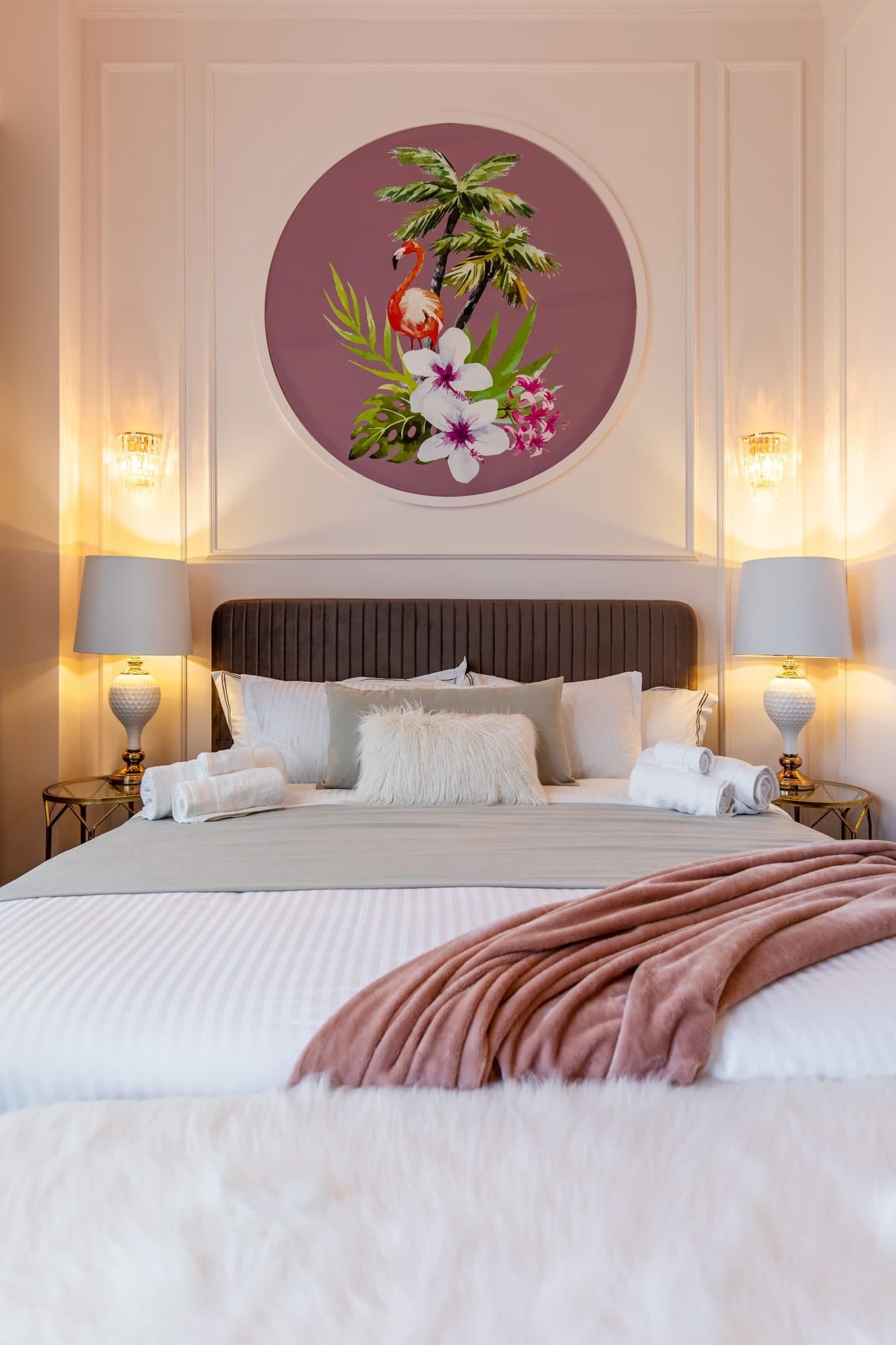 A neatly made bed with white linens and a fluffy white throw, two bedside lamps, and a wall-mounted circular artwork depicting tropical flowers and a flamingo.
