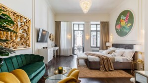 A hotel room with a bed, a green sofa, a yellow armchair, a glass coffee table, and a large decorative wall piece.