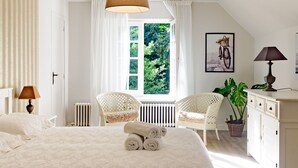 Ein Schlafzimmer mit einem großen Bett, einem Fenster mit Blick auf Grünflächen, einem Korbstuhl, einer Topfpflanze und einer Kommode.