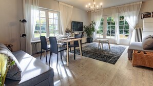 Ein modernes Wohnzimmer mit einer Couch, einem Esstisch, Stühlen, einem Fernseher und einem großen Fenster mit Vorhängen.