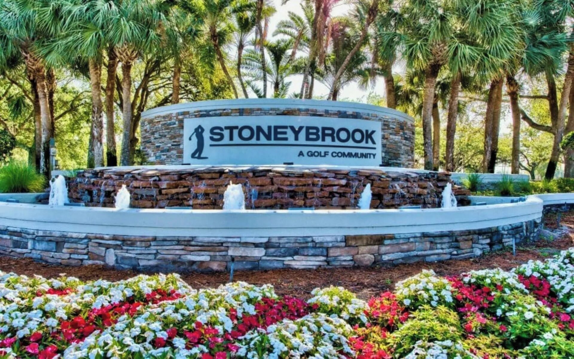 Une fontaine en pierre avec des jets d’eau, située dans un espace paysager agrémenté de fleurs aux couleurs vives, entourée de palmiers et d’un panneau indiquant : « Stoneybrook, un quartier résidentiel avec terrain de golf ».