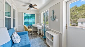 Une pièce de taille modeste, avec un canapé bleu, un bureau, un ventilateur de plafond et une vue sur l’extérieur.