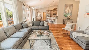 A spacious living room with a large sectional sofa, a coffee table, and a dining area with a kitchen in the background. - Gorgeous, Spacious and Well Lit Hot Tub Home with Large Deck for Entertaining. (Rockaway Beach)