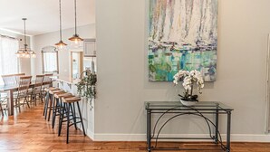 A modern kitchen with wooden flooring, pendant lighting, a dining area with bar stools, and a large abstract painting on the wall. - Gorgeous, Spacious and Well Lit Hot Tub Home with Large Deck for Entertaining. (Rockaway Beach)