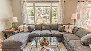 A living room with a sectional sofa, a glass coffee table, and a view of a backyard with a patio and trees. - Gorgeous, Spacious and Well Lit Hot Tub Home with Large Deck for Entertaining. (Rockaway Beach)