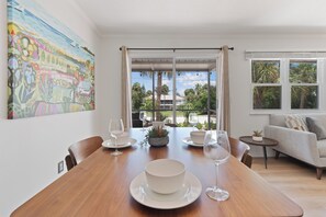 A dining area with a wooden table set for four, a sofa, and a view of a garden through large windows.