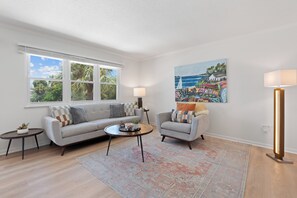 A living room with a sofa, a coffee table, a side table, a floor lamp, a painting, and a window with a view of trees and sky.