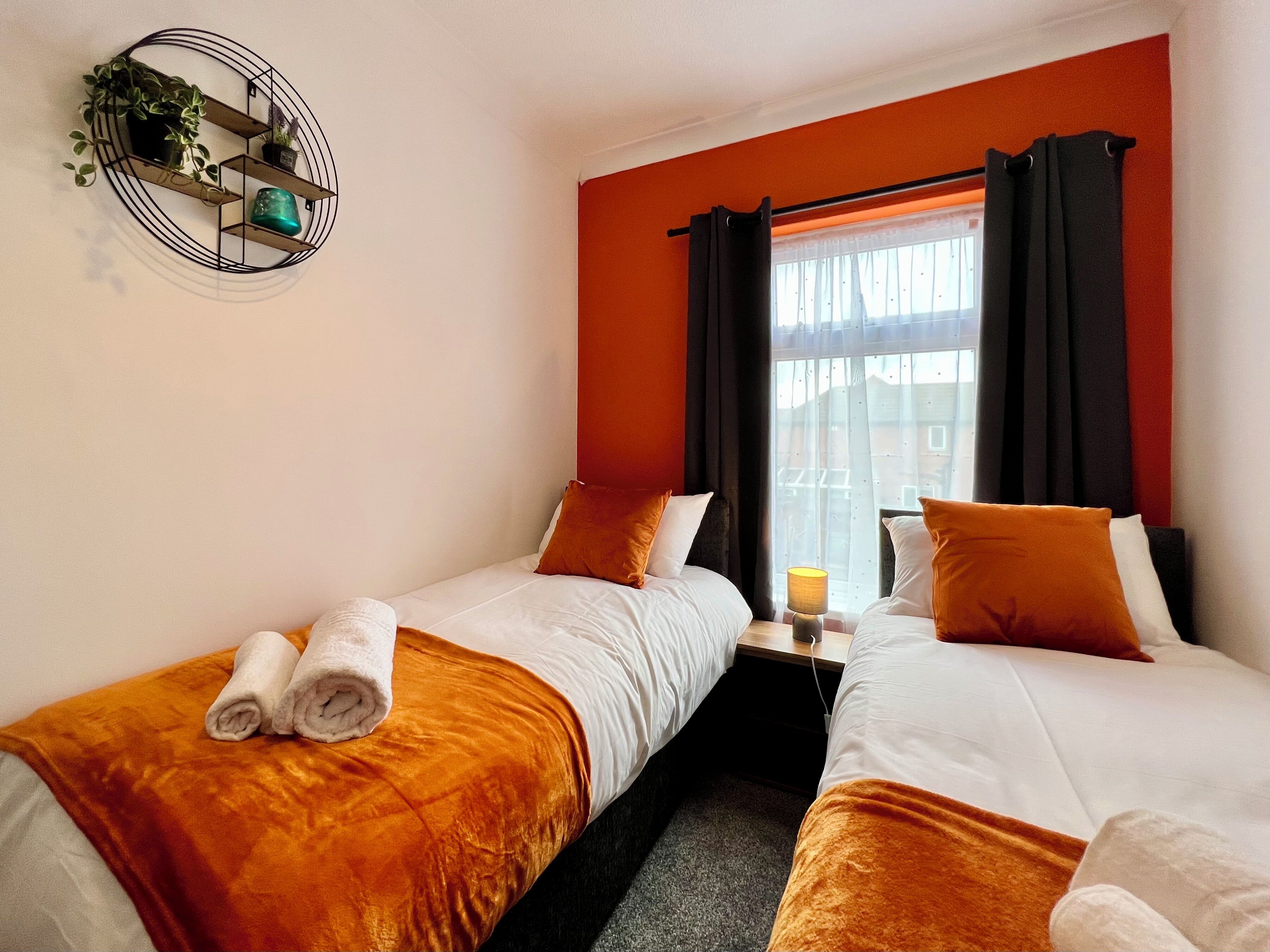 Una habitación de hotel con dos camas, ropa de cama naranja, una mesita con una lámpara y una ventana con cortinas.