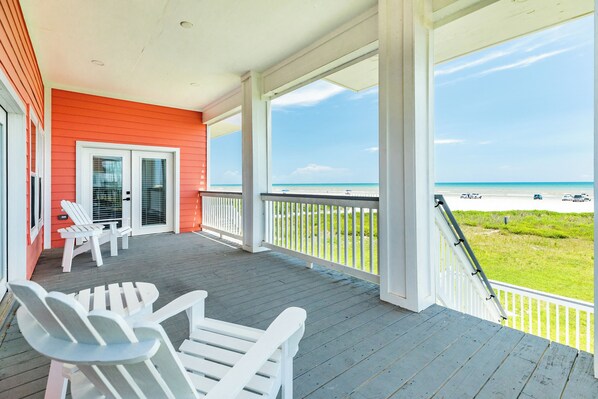 A covered porch with white Adirondack chairs, a view of the beach, and a red building in the background.