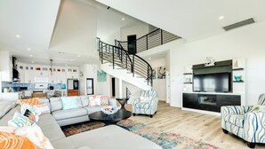 Un vaste espace de vie comprenant un grand canapé, une table basse et un tapis à motifs. Un escalier avec une rampe noire mène à un niveau supérieur.