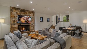 A modern living room with a stone fireplace, a flat-screen TV, a dining area with a table set for six, and a staircase leading to an upper level. - Perfect Perch With All the Views | Radiance Retreat (Vail)