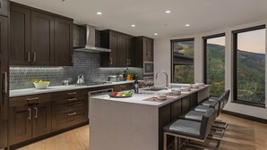 A modern kitchen with dark wood cabinets, a central island with seating, and large windows offering a view of the outdoors. - Perfect Perch With All the Views | Radiance Retreat (Vail)