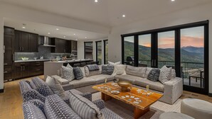A modern living room with a large sectional sofa, a wooden coffee table with playing cards, and a view of mountains through large windows. - Perfect Perch With All the Views | Radiance Retreat (Vail)