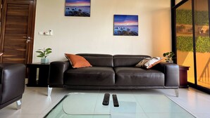 En stue med en lædersofa, to billeder hængt på væggen, et glashøjt sofabord og en skydedør i glas, der fører ud til en balkon.