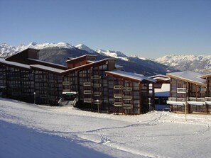 Une station de ski enneigée, avec des bâtiments en bois, un ciel dégagé et des montagnes enneigées en arrière-plan.