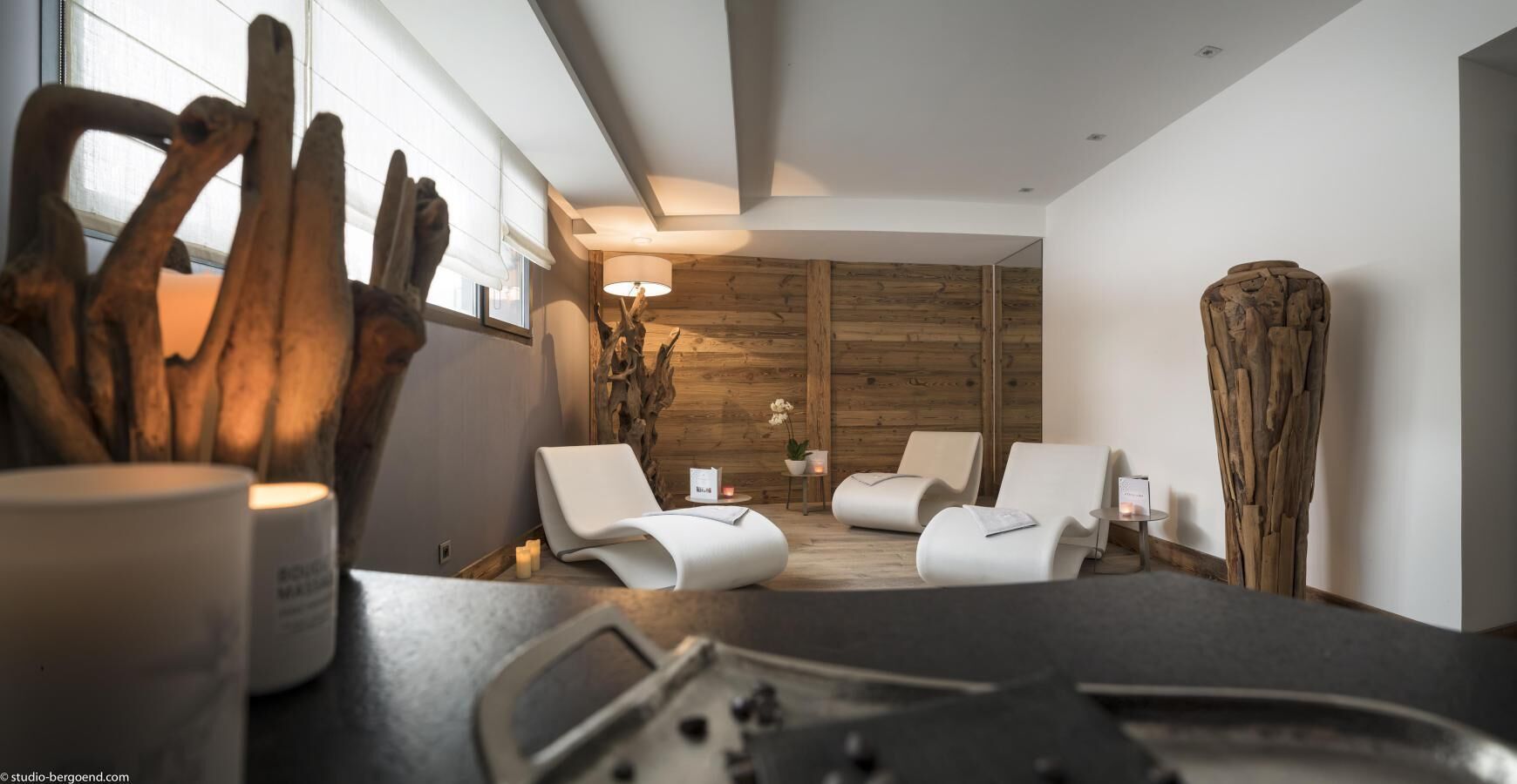 A modern spa room with a wooden wall, two white lounge chairs, a small table with candles, and a large wooden sculpture.