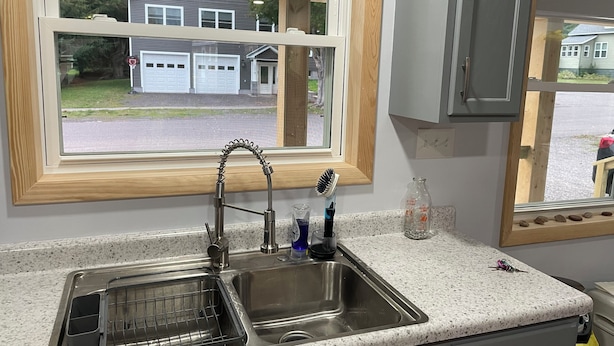 Una cocina con un lavamanos doble, una ventana con vistas a casas y una encimera con un escurreplatos y una botella.