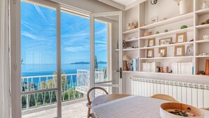 Une chambre avec un balcon donnant sur la mer, des étagères blanches ornées d’objets décoratifs et une table recouverte d’une nappe tissée.