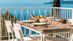 Une table en bois, des chaises blanches, un saladier rempli de fruits et une vue sur la mer.