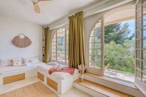 Une chambre à coucher avec un lit, une fenêtre avec des rideaux, un ventilateur de plafond et une vue sur la verdure à l’extérieur.