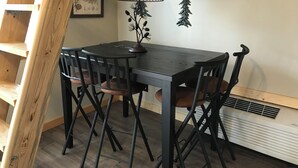 A dining area with a dark wooden table, black chairs, a yellow lamp, a pine tree decoration, and a 'Welcome to Cabin' sign. - Judy's Place - 1BR, 1 Bath w/ Q, K, and Twin bed. Full kitchen. (Putney)
