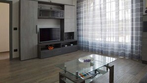 Una sala moderna con una mesa de centro de vidrio, un mueble de entretenimiento gris y cortinas translúcidas.