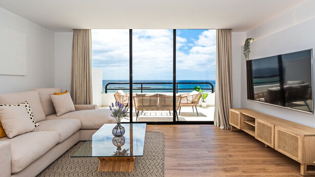 Un salon moderne avec un canapé, une table basse et un téléviseur, donnant sur un balcon avec vue sur l’océan.