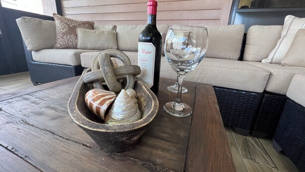 Ein Holztisch mit einer Weinflasche und einem Glas, eine Schale voll Meeresschalen und ein Korbsessel mit Kissen.