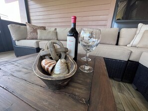 Ein Holztisch mit einer Weinflasche und einem Glas, eine Schale voll Meeresschalen und ein Korbsessel mit Kissen.