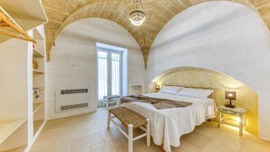 Une chambre à coucher avec un plafond voûté, un lit avec une tête de lit, un banc, une table de chevet avec une lampe et une fenêtre donnant sur un beau paysage.