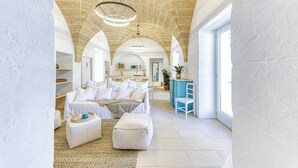 Une pièce spacieuse avec un plafond en pierre, un grand lit avec une literie blanche, une chaise bleue et un coin cuisine à l’arrière-plan.