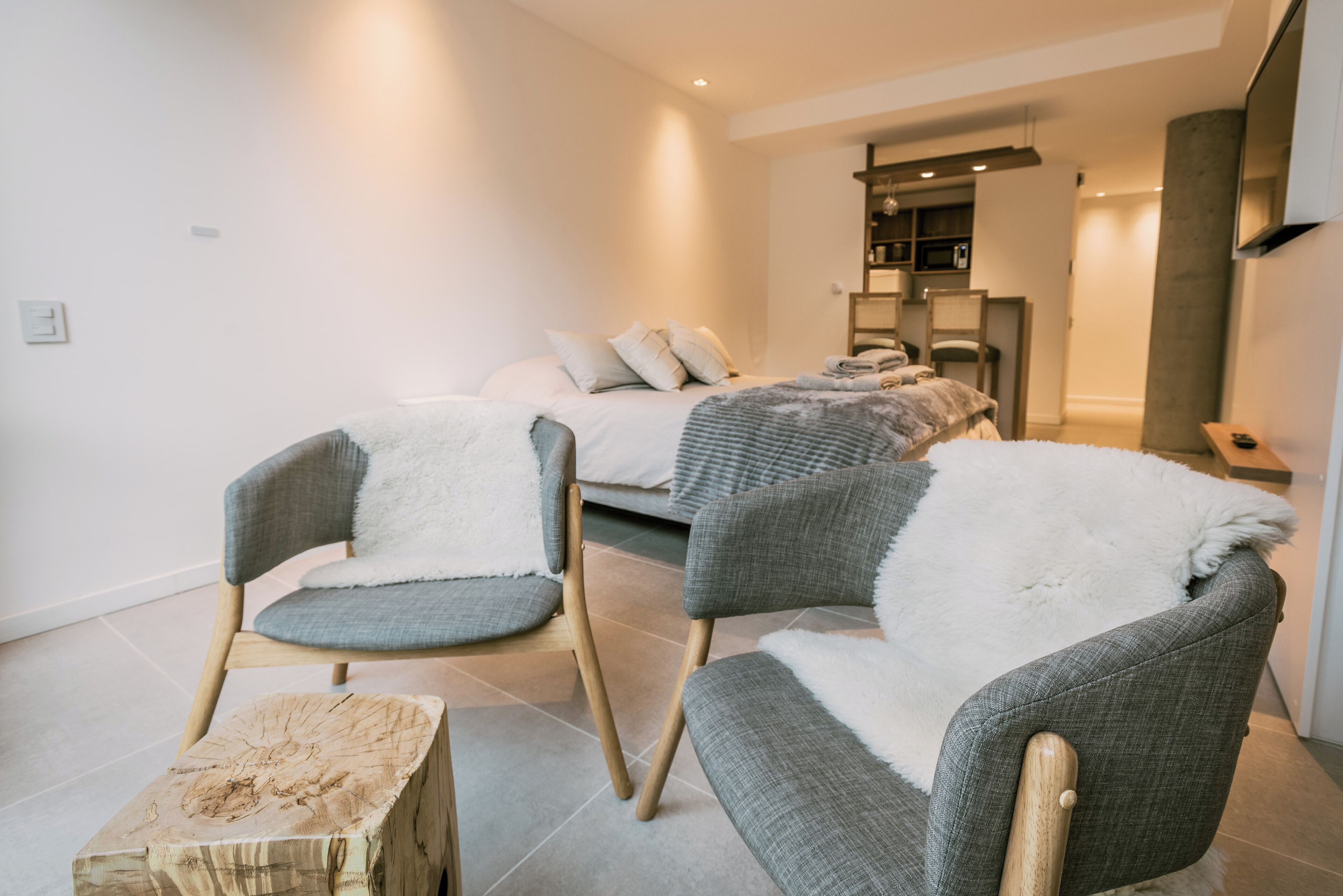 A modern bedroom with a bed, two grey upholstered chairs, a small wooden stool, and a built-in wardrobe.