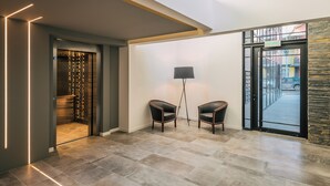 Un vestíbulo moderno con un ascensor de vidrio, dos sillones de cuero, una lámpara de pie y vistas al exterior a través de una puerta de cristal.