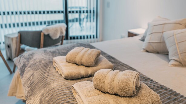 Una cama bien hecha con toallas enrolladas, una silla con una manta y un ventanal con persianas.