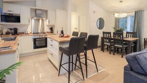 A modern kitchen with white cabinets, a central island with bar stools, a dining area with a wooden table and chairs, and a living area with a sofa. - Unique Luxury Lodge (Windermere)
