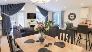 A modern living room with a dining area, featuring a wooden table, heart-shaped placemats, a vase with flowers, and a large wall clock. - Unique Luxury Lodge (Windermere)