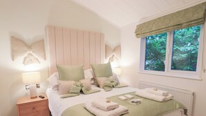 A neatly arranged bedroom with a bed, bedside lamps, a nightstand, and a window with a view of greenery. - Unique Luxury Lodge (Windermere)