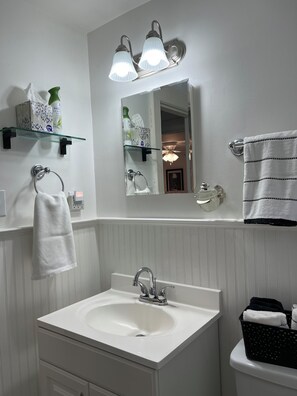 Een badkamer met een witte wastafel, een spiegel en een handdoekrek. Boven de spiegel zijn planken met toiletartikelen.
