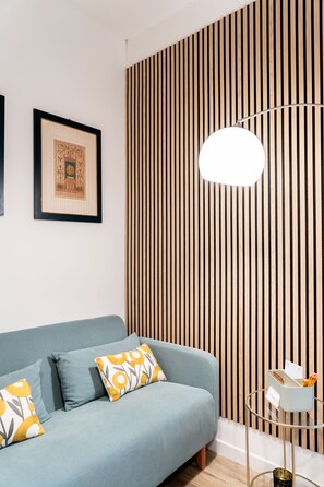 A modern living room with a blue sofa, patterned cushions, a framed picture, a vertical wooden panel, and a glass table with a white base. - Beautiful, modern three-room apartment for 4 people, with 2 bedrooms, 2 bathrooms, a laundry room, and a parking space (Perugia)