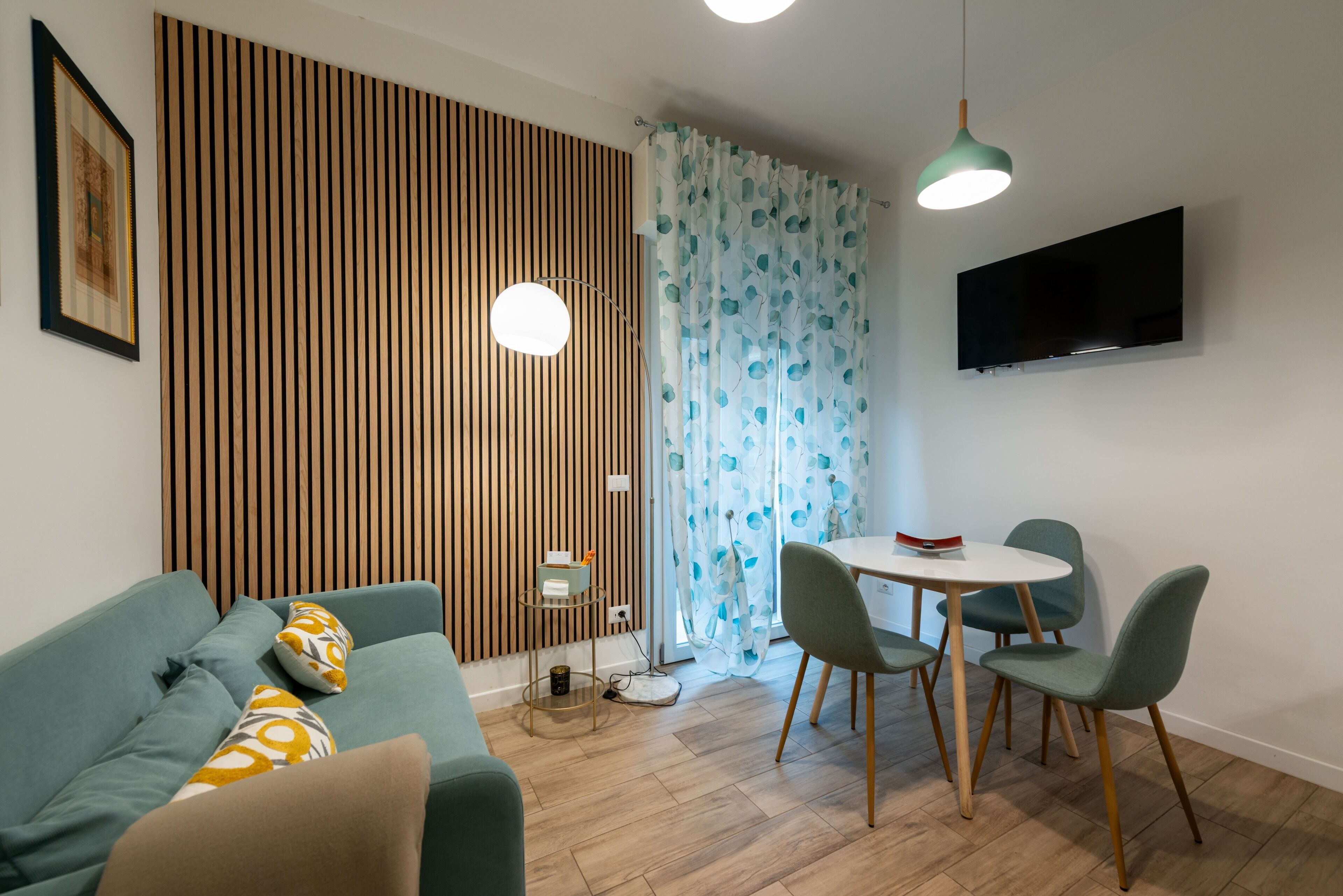 Ein modernes Wohnzimmer mit einer Couch, einem Esstisch mit Stühlen, einem an der Wand montierten Fernseher und einem gemusterten Vorhang.