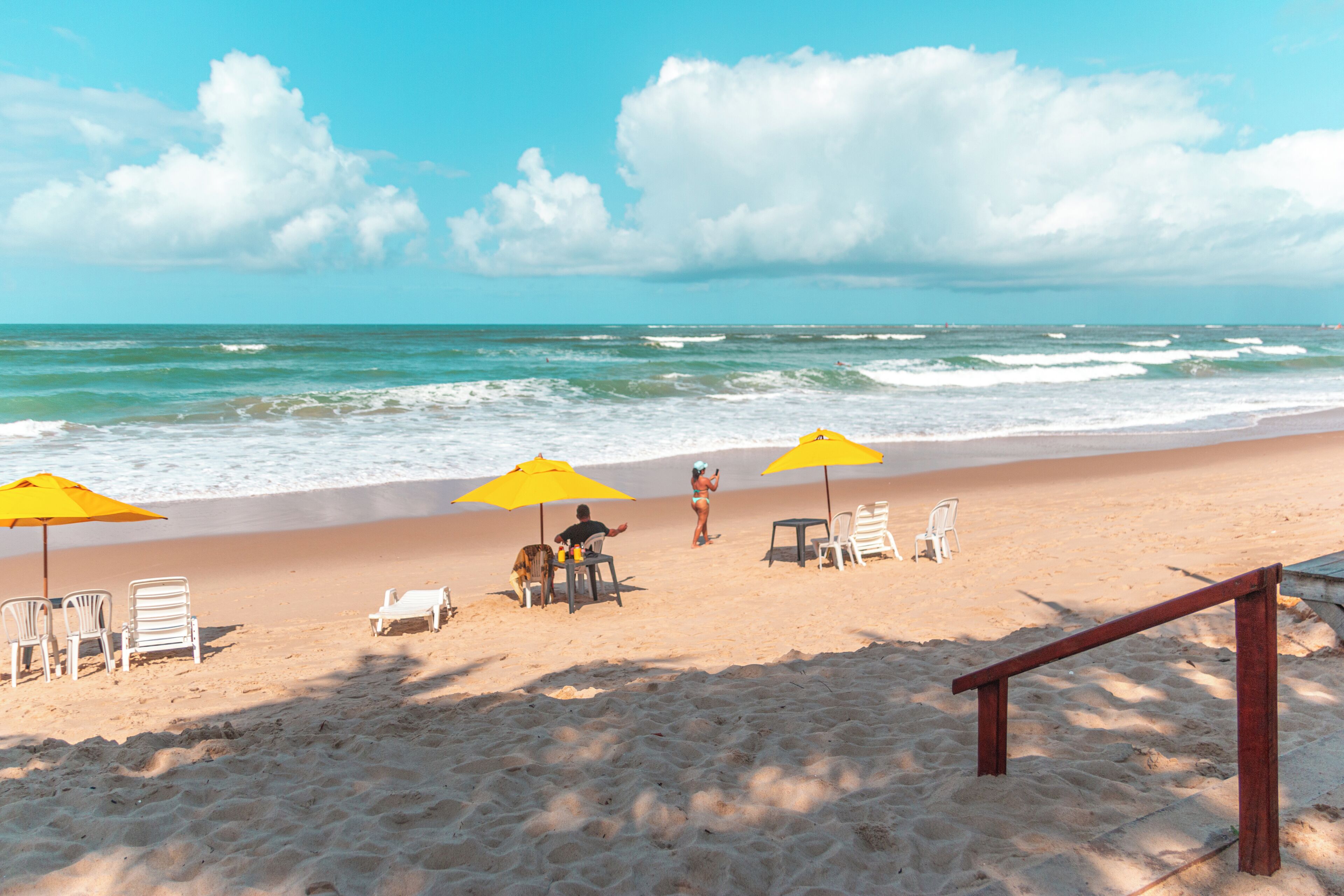 Ein Strand mit gelben Sonnenschirmen, weißen Stühlen und einer Holzbank.