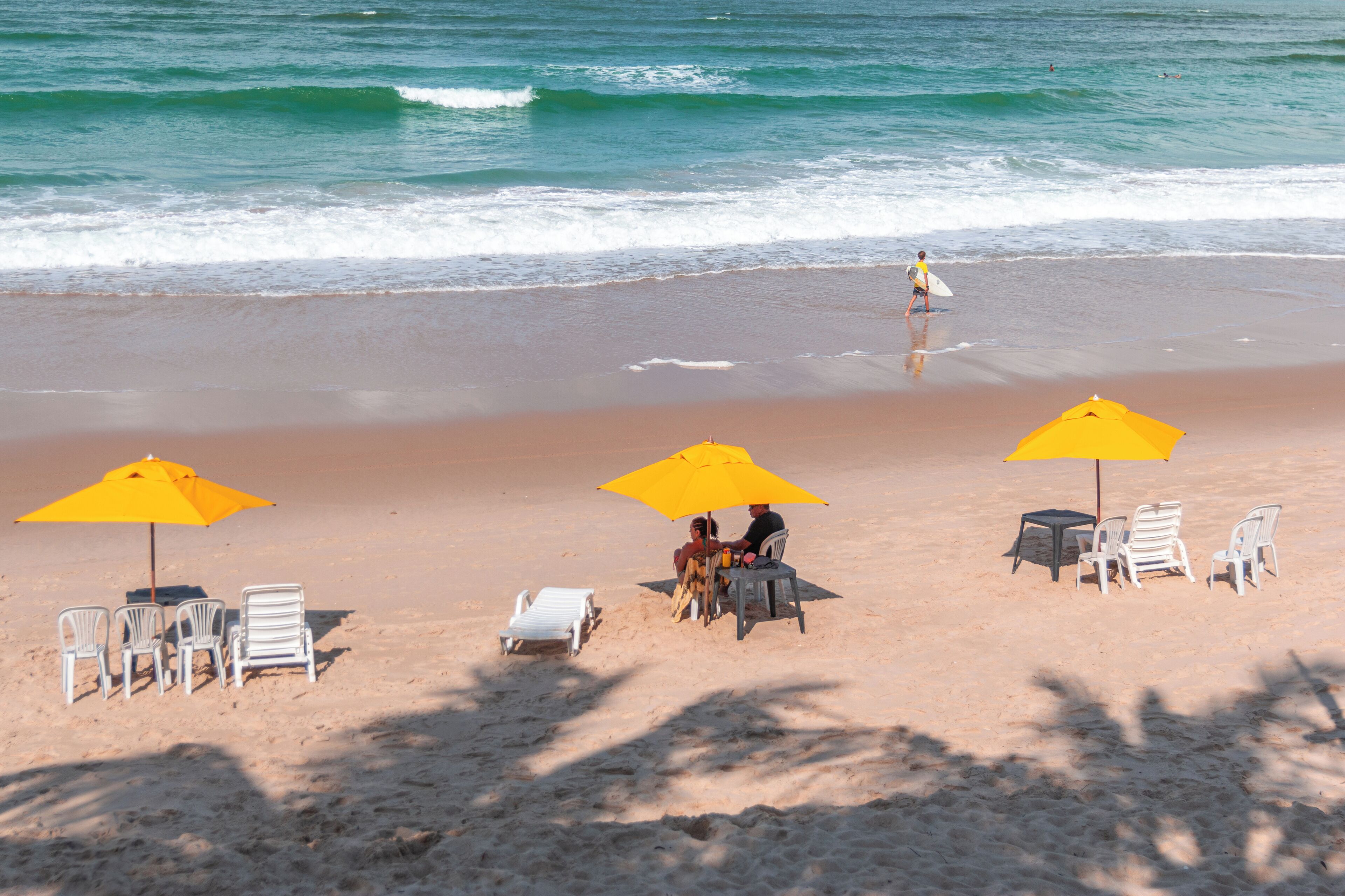 Une plage avec des chaises longues blanches et des parasols jaunes, une table, et une personne qui se promène avec une planche de surf.