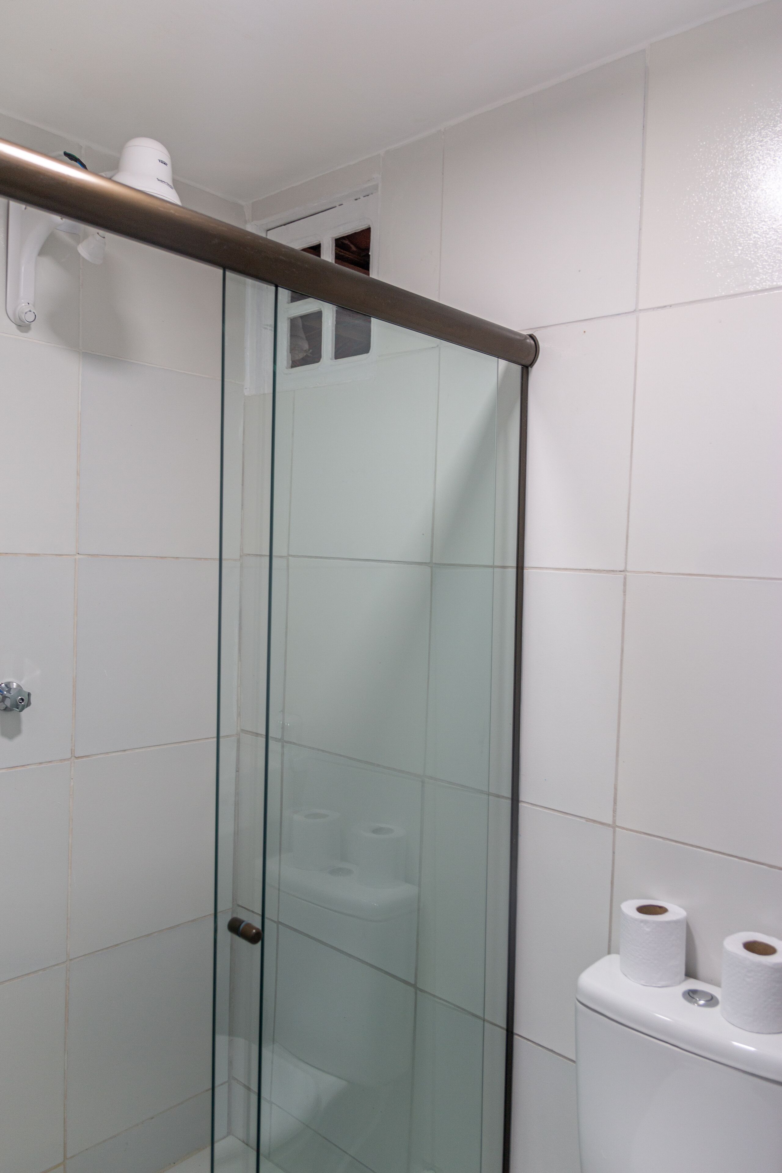 A glass-enclosed shower stall with a metal handle, white tiled walls, and a toilet with two rolls of toilet paper.