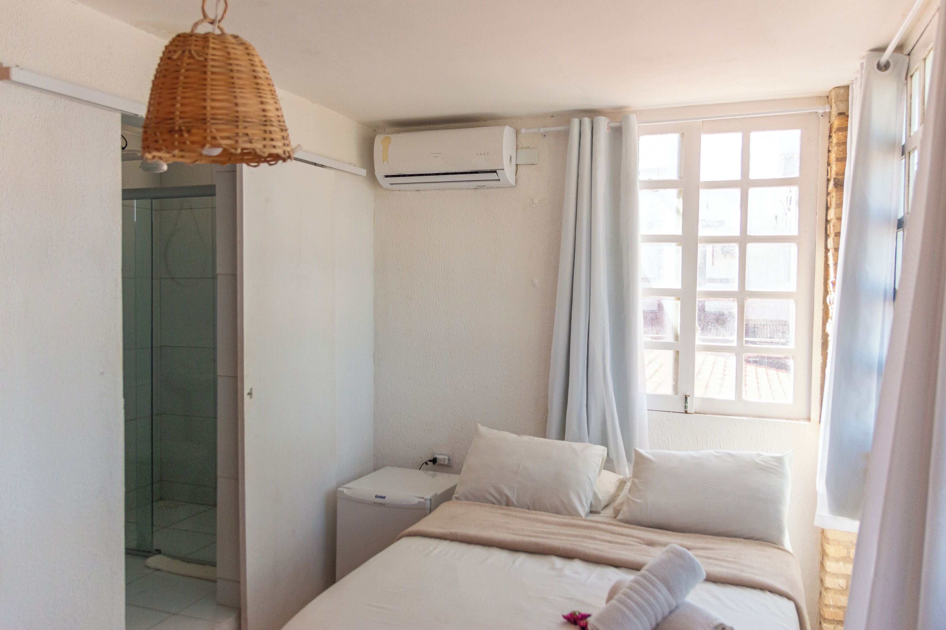 Ein Schlafzimmer mit Bett, Nachttisch, Wandairconditioner, Fenster mit Vorhängen und einer Duschkabine.