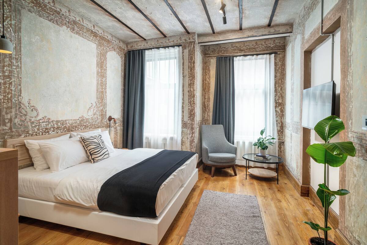 A bedroom with a large bed, a chair, a small table, and a potted plant.