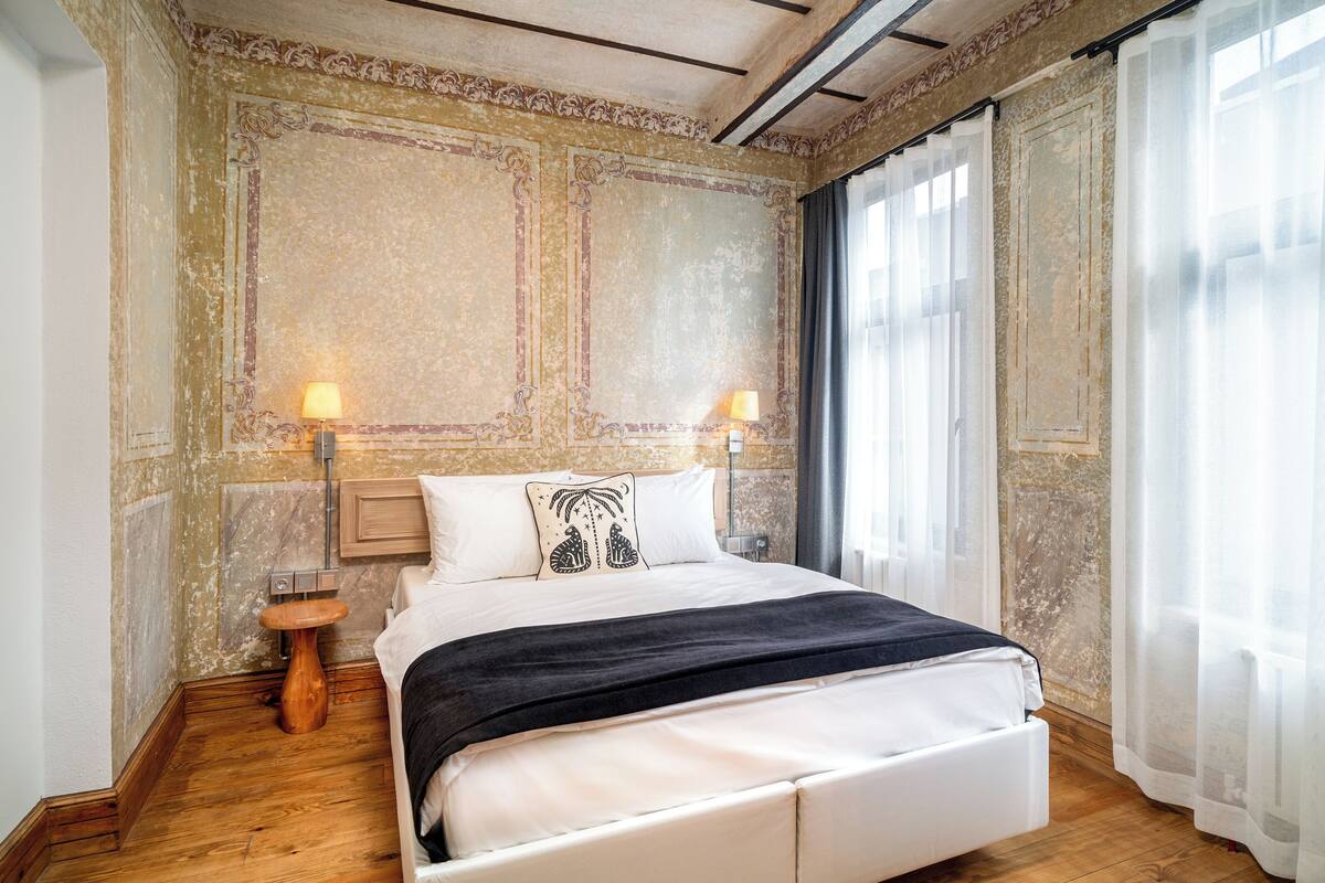 A bedroom with a bed, two bedside lamps, a wooden nightstand, and a large window with sheer curtains.