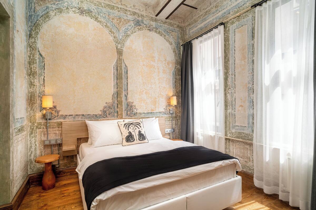 A bedroom with a large bed, two bedside lamps, a wooden stool, and a view of the outside through sheer curtains.